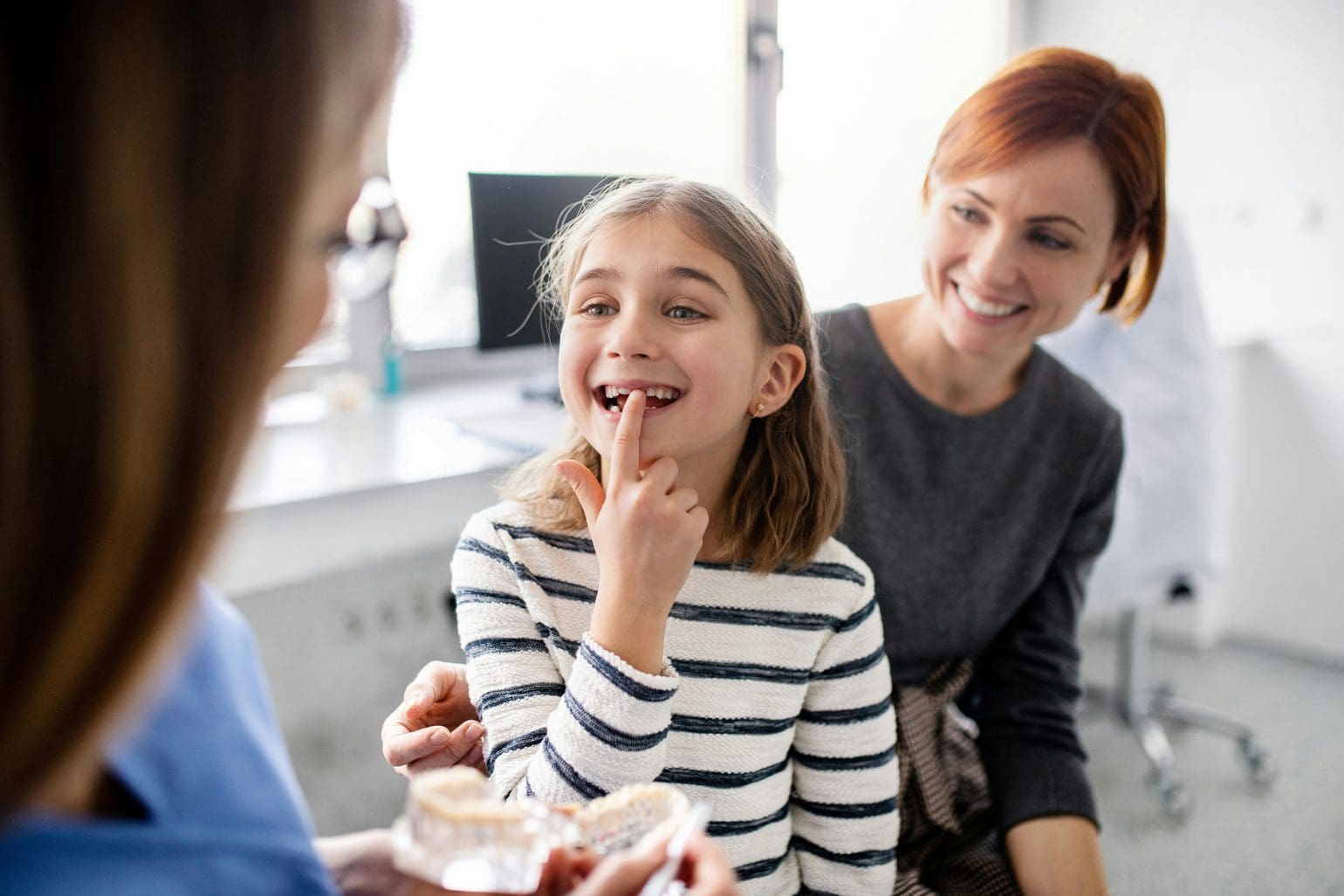 little girl shows her tooth to the dentist while her mother sits behind her, smiling